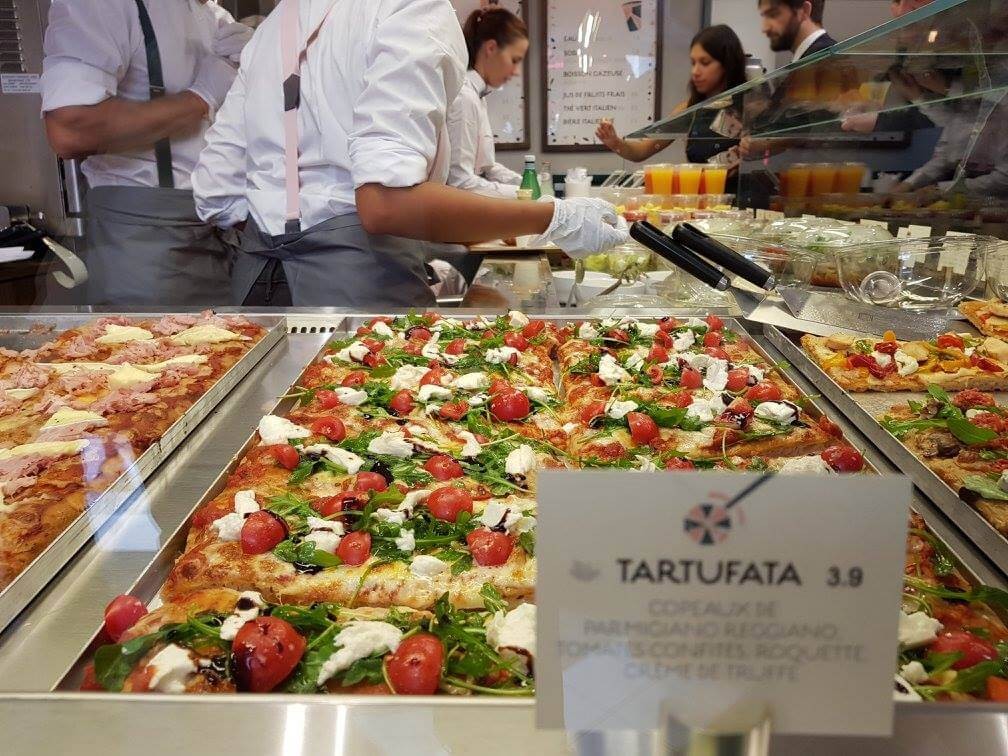 Plaques de pizza al taglio exposées sous vitrage au comptoir, équipe en préparation