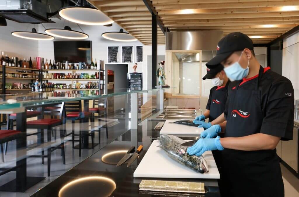 Sushimans préparant des makis sur un comptoir réfrigéré face à la salle d'un kiosque à sushis