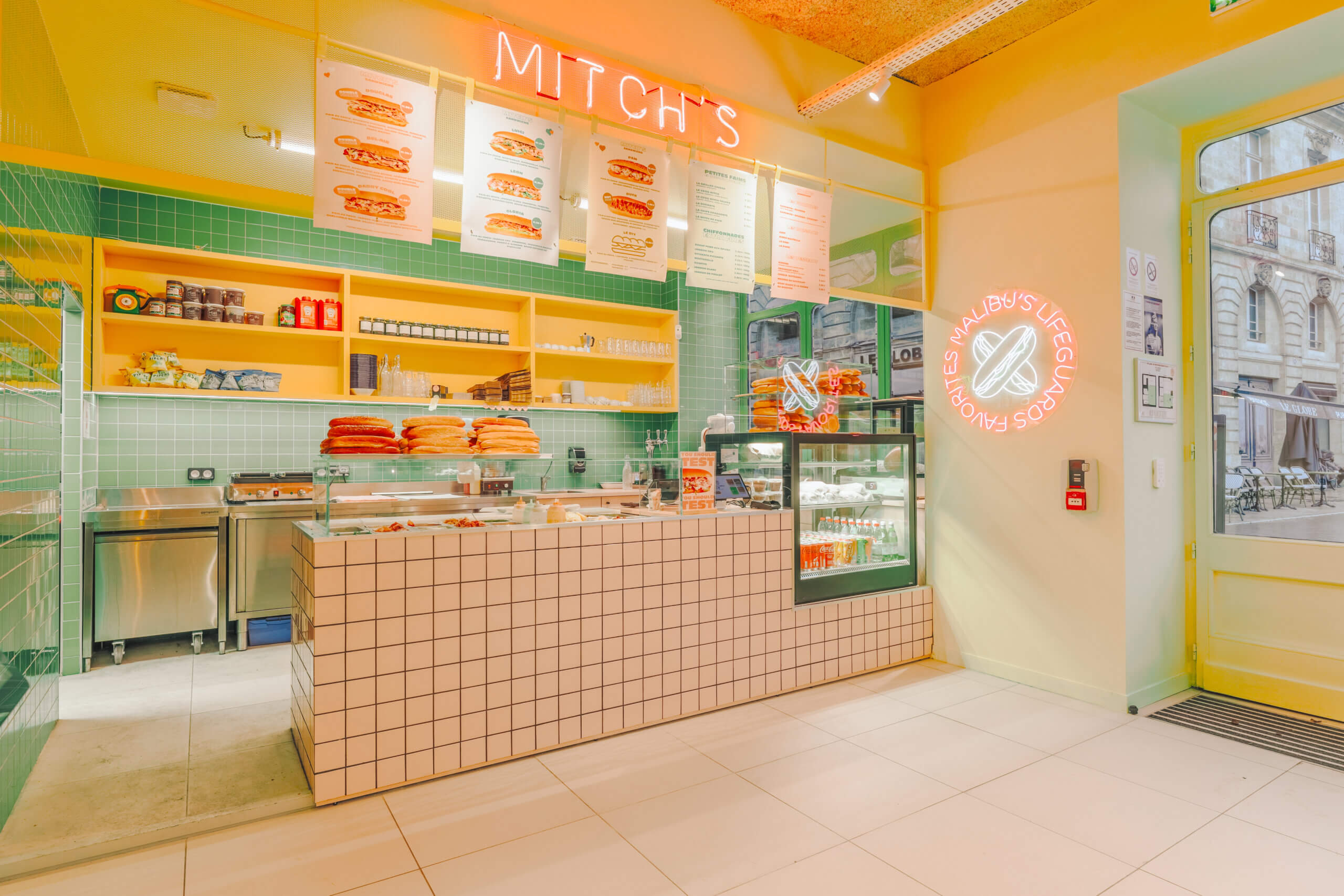 Salle de la sandwicherie Mitch's avec comptoir carrelé, enseigne néon et vitrine réfrigérée