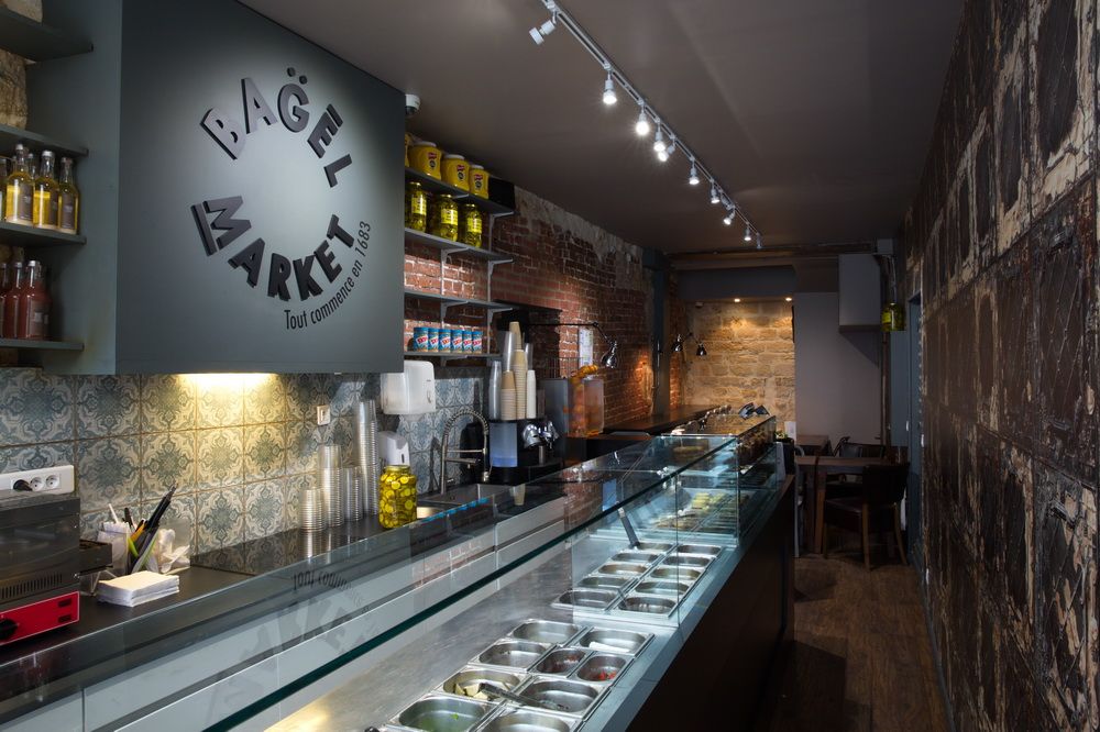 Bagel store : long comptoir vitré avec ingrédients en bacs GN réfrigérés, murs en brique et carreaux de ciment