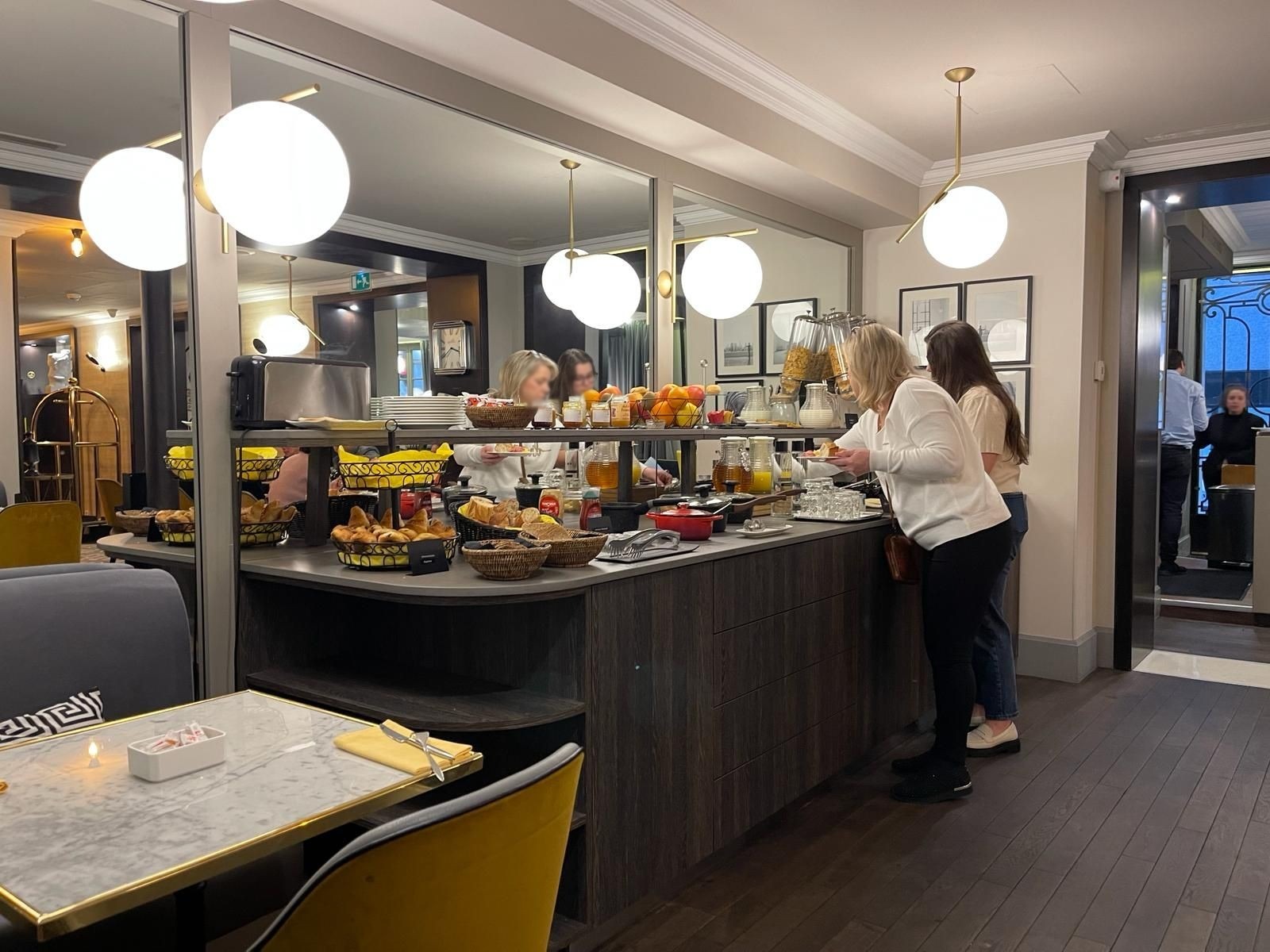 Clients au buffet de petit-déjeuner d’un hôtel, comptoir bois foncé et suspensions globes