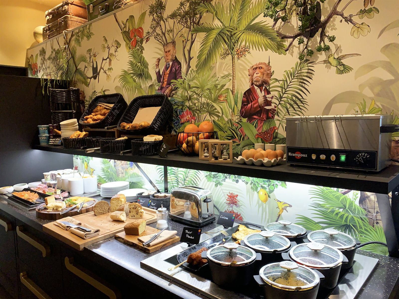 Buffet de petit-déjeuner d’hôtel : viennoiseries, fromages, cocottes chaudes et toasteur devant un décor végétal peint