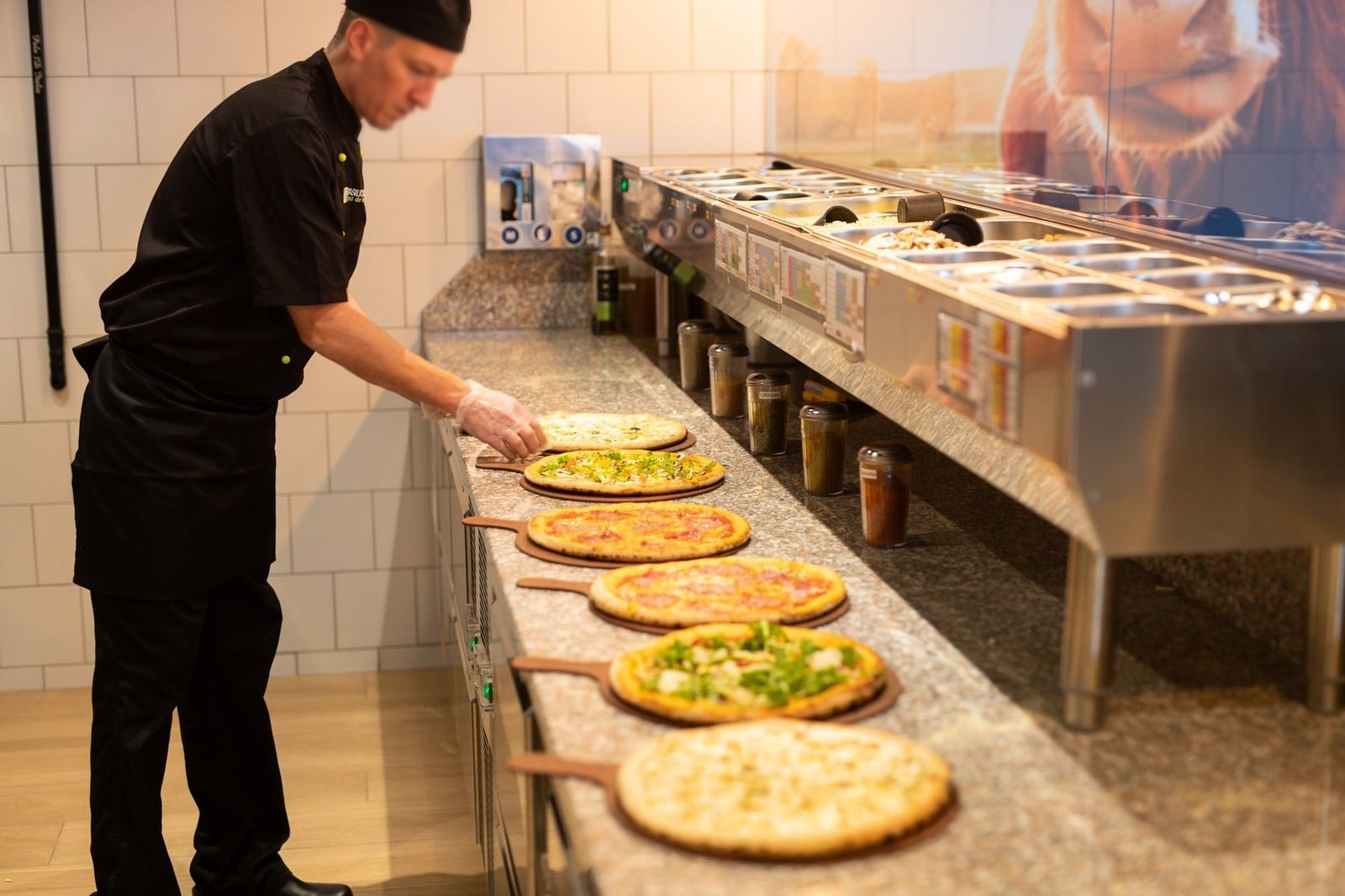 Pizzaiolo garnissant des pizzas sur une table de préparation avec bacs à toppings réfrigérés