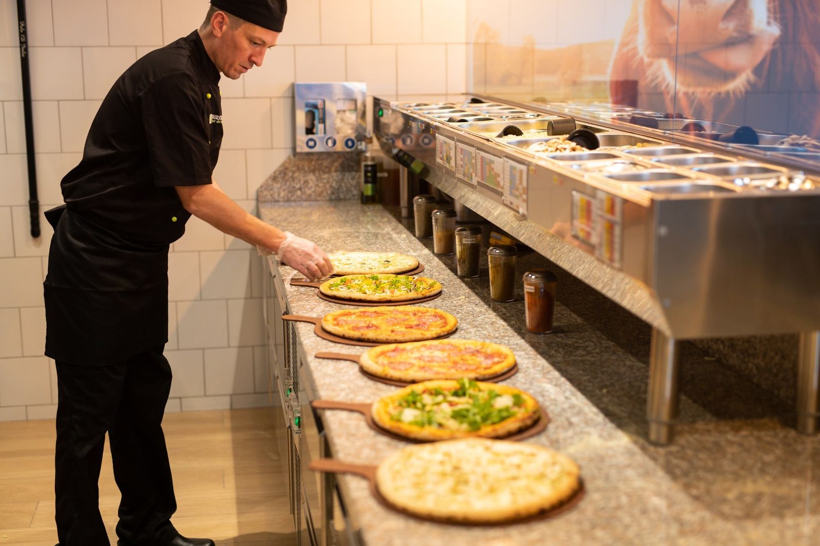 Pizzaiolo garnissant des pizzas sur une table de préparation avec bacs à toppings réfrigérés