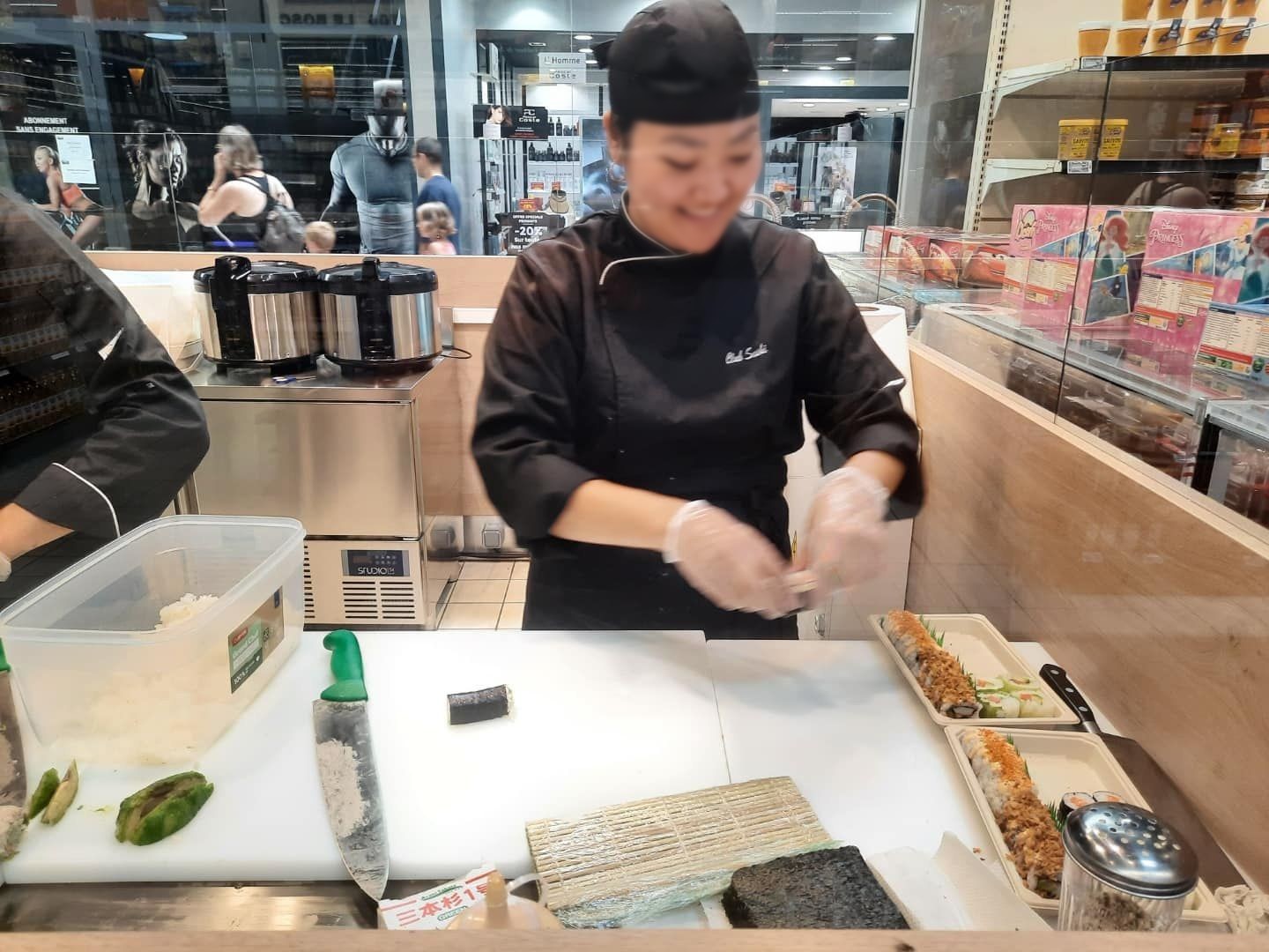 Sushiman au poste de préparation avec planche de découpe, cuiseurs à riz et vitrine à proximité