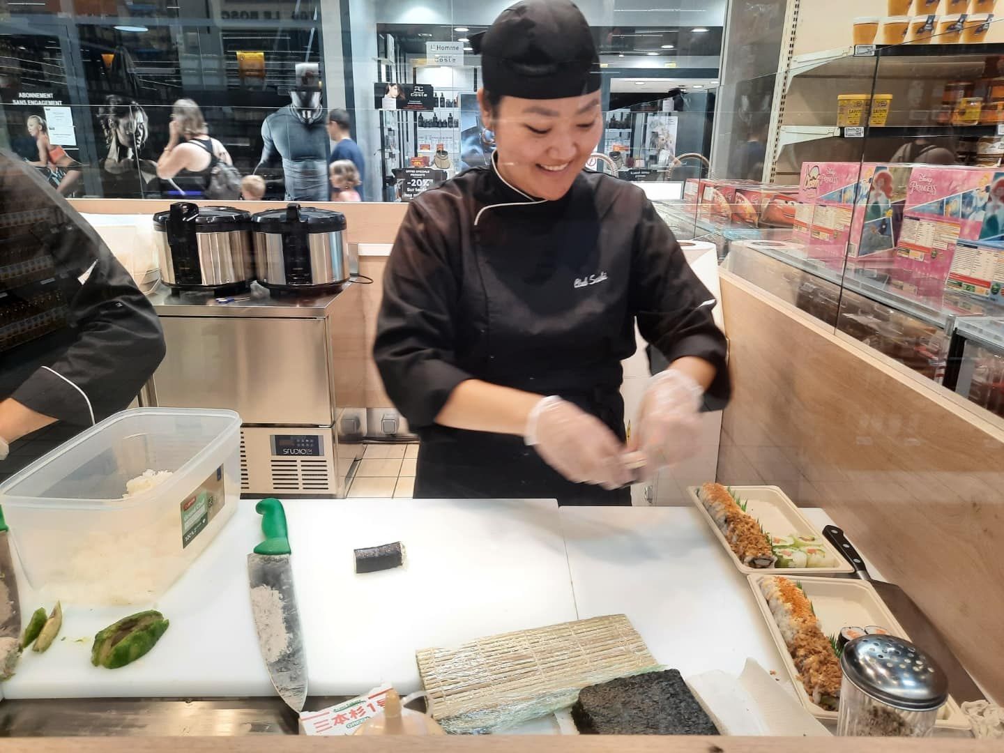 Sushiman au poste de préparation avec planche de découpe, cuiseurs à riz et vitrine à proximité