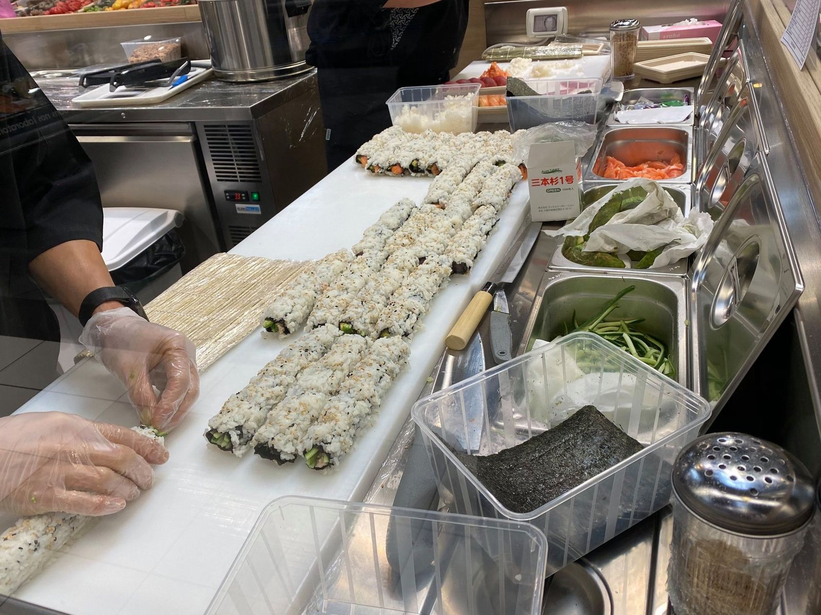 Préparation de makis sur une planche blanche avec bacs d’ingrédients réfrigérés et feuilles d’algue