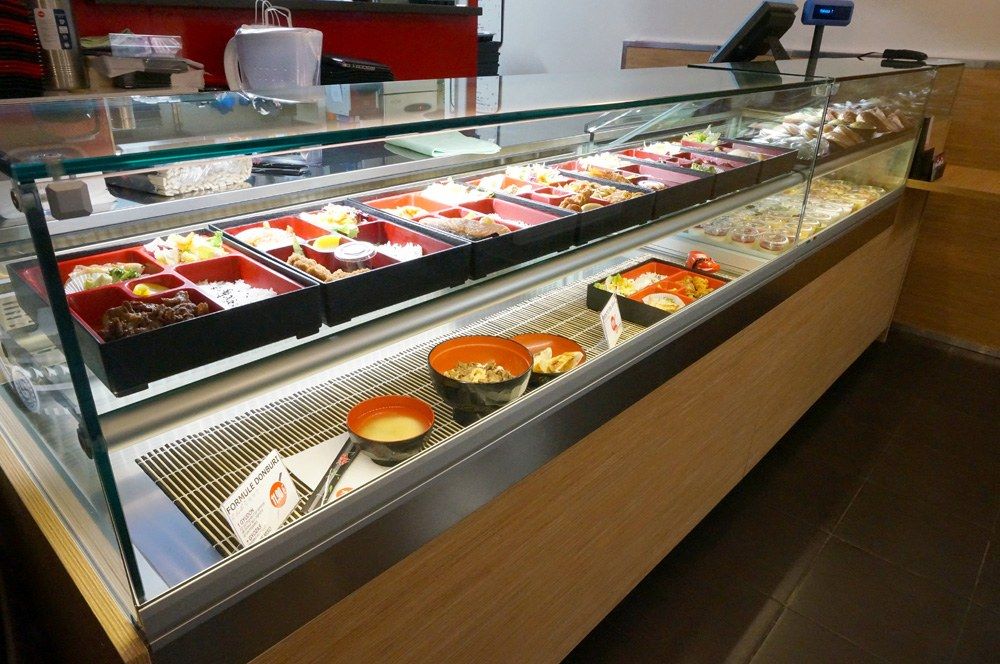 Vitrine réfrigérée de restaurant japonais présentant bentos, soupes et plats sur tapis de bambou