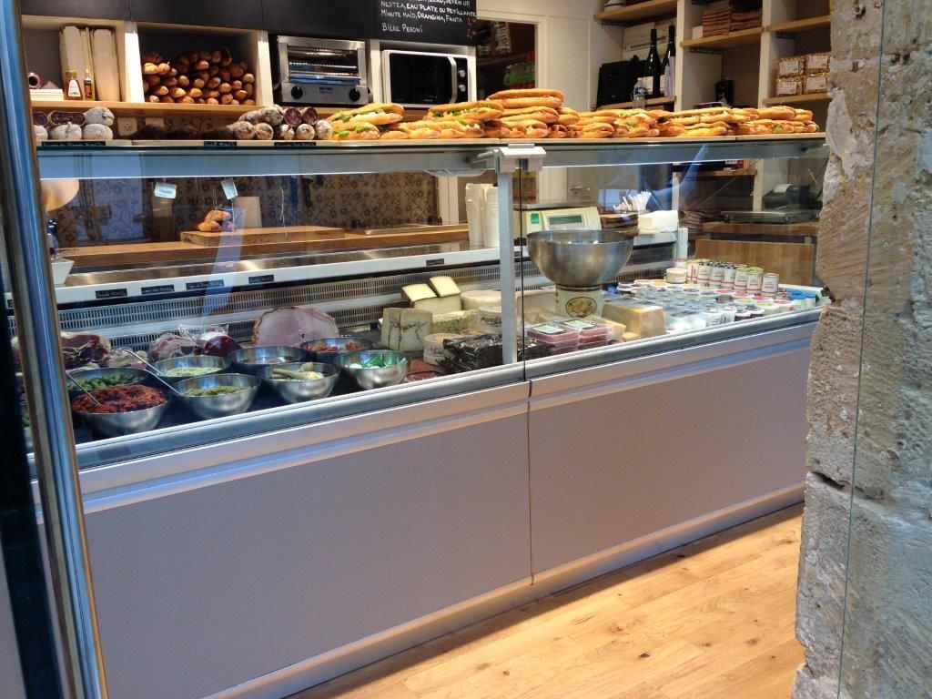 Vitrine réfrigérée traiteur garnie de charcuteries, salades et fromages, sandwichs présentés au-dessus